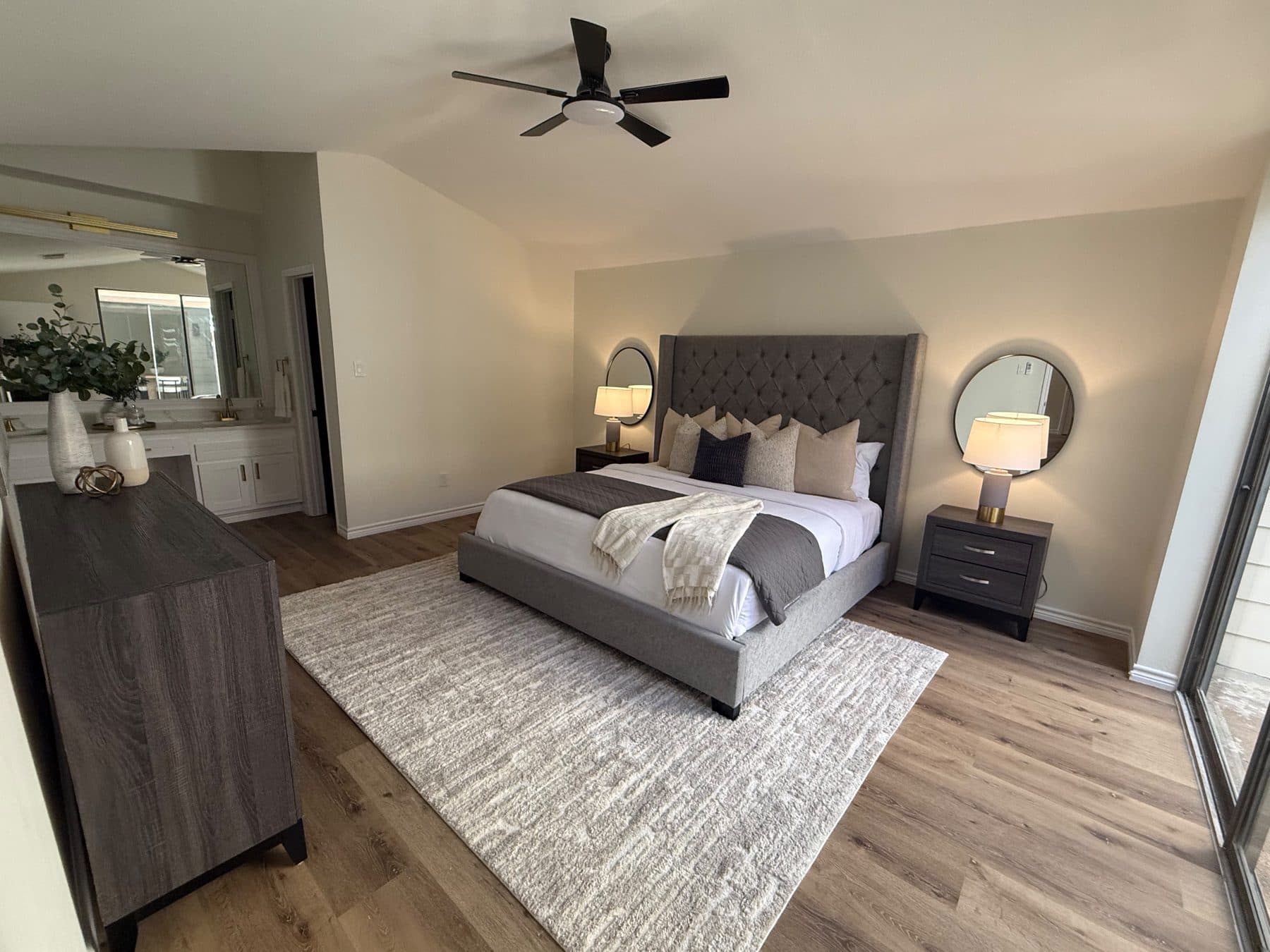 Primary bedroom from the window side with the upholstered bed and round mirror, staged for listing and finished by the Reborn group